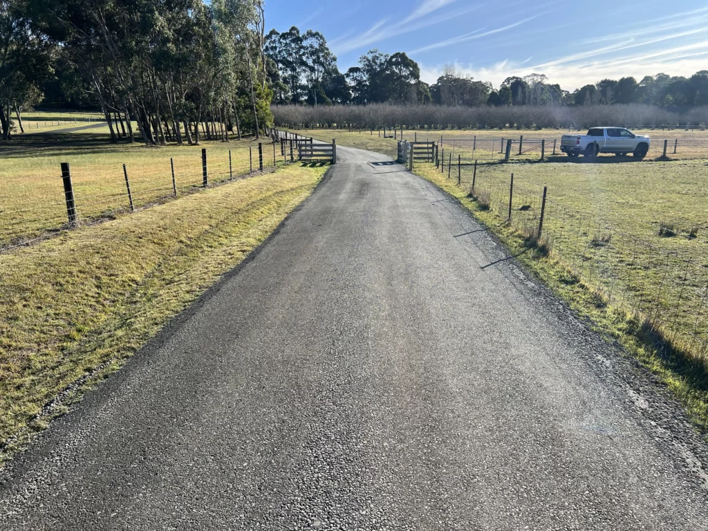 Sealed asphalt driveway running through a rural property with a white vehicle parked near a fence.